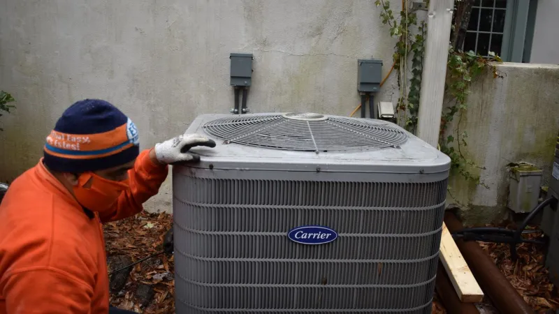 Technician in orange jacket inspecting a Carrier air conditioning unit outside a house with ivy on the wall.