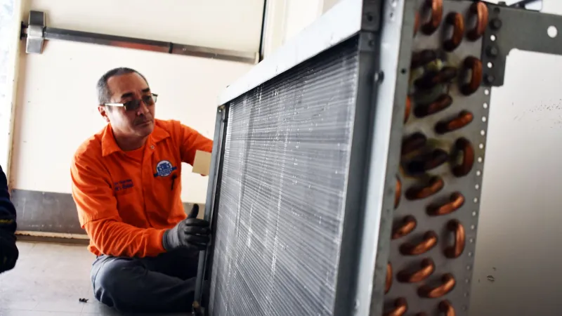 Technician in orange uniform inspecting a large HVAC heat exchanger coil indoors on the floor.