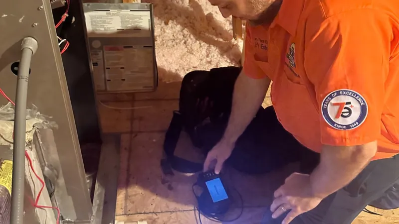 Estes Services Technician wearing an orange uniform shirt and headlamp inspecting HVAC system with electronic device and power tools in attic