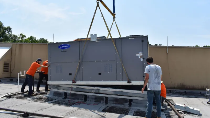 Technicians installing a Carrier HVAC unit on a flat rooftop using a crane on a sunny day.