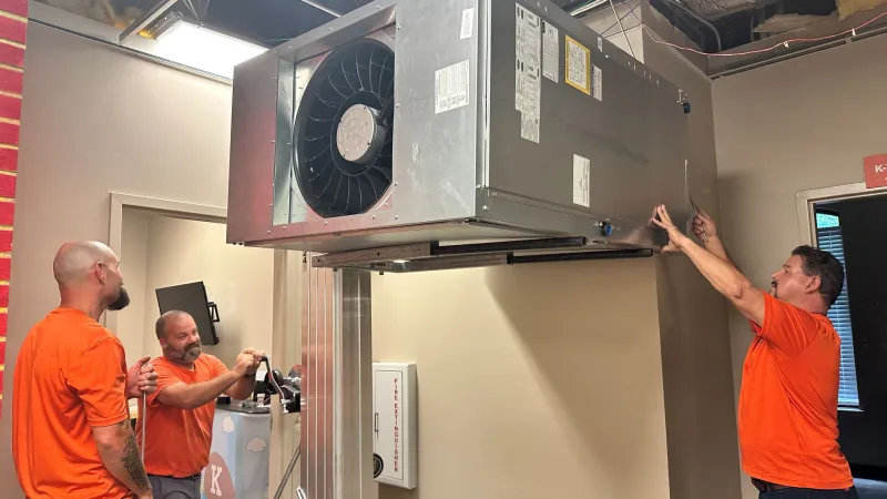 Three workers in orange shirts installing a large HVAC unit inside a building under construction.