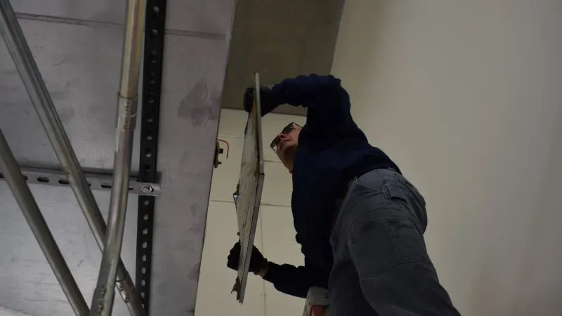 Man wearing gloves installs ceiling panel while standing on ladder in an indoor construction site.