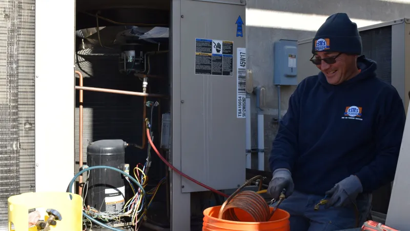 Technician wearing a beanie and gloves working on HVAC unit with tools and orange bucket outdoors