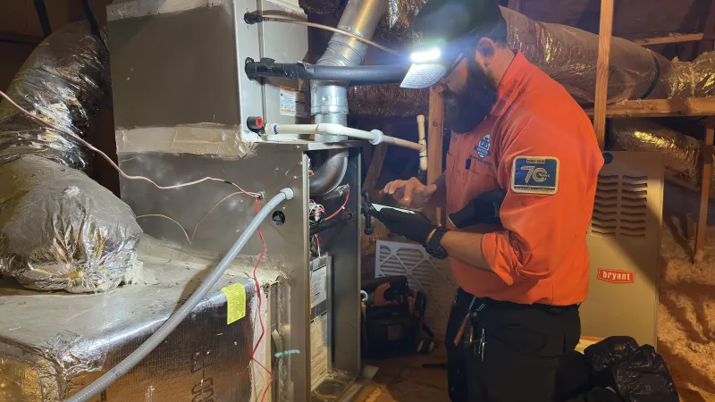 Technician inspecting and repairing HVAC system in attic wearing orange shirt and headlamp in dimly lit space