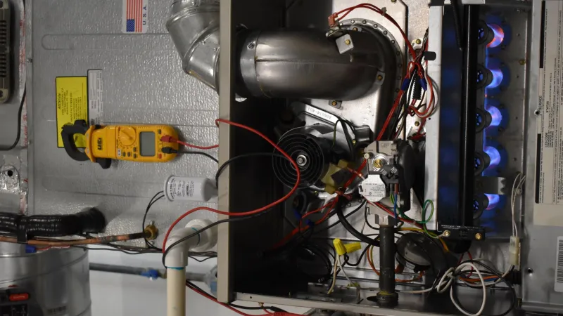 Open furnace with visible blue flames, wires, pipes, and a digital multimeter for diagnostics and repair work