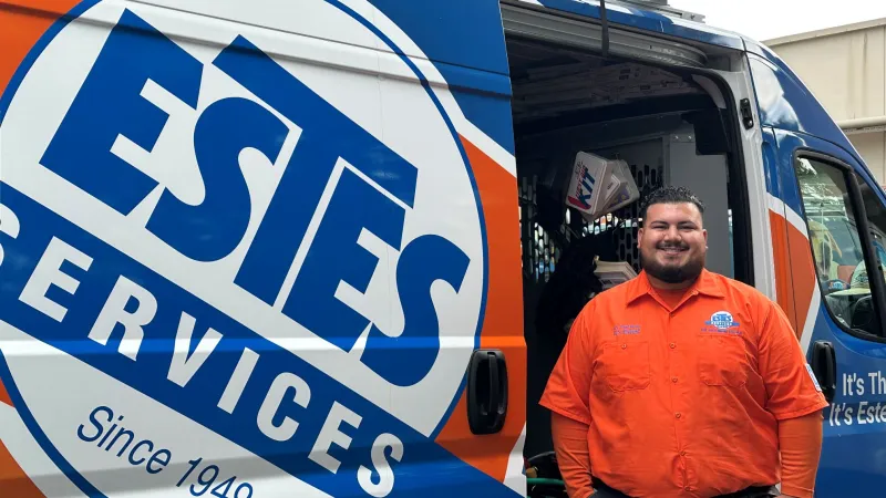 Smiling Estes Services technician in orange shirt standing next to a blue and orange company service van with ladder.