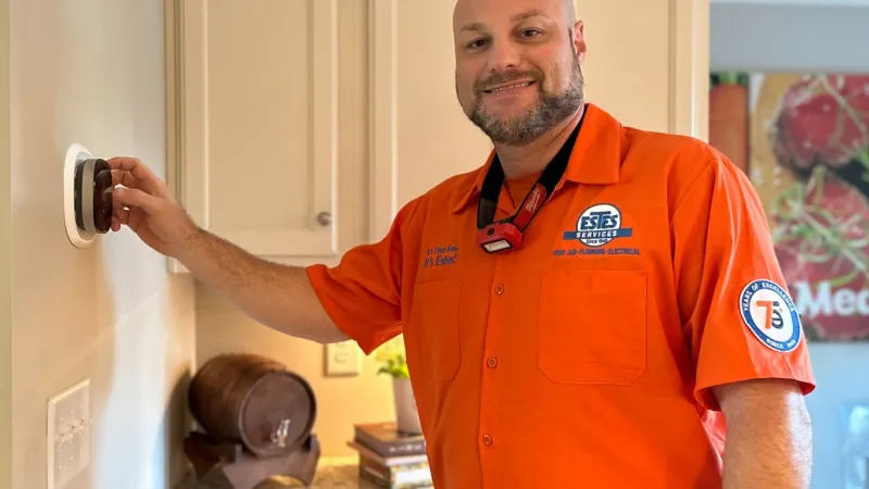 Estes Services Technician in orange uniform adjusting a thermostat on a kitchen wall with cabinets in the background