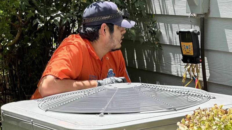 Technician in orange shirt inspecting a Carrier air conditioning unit outside a house on a sunny day.