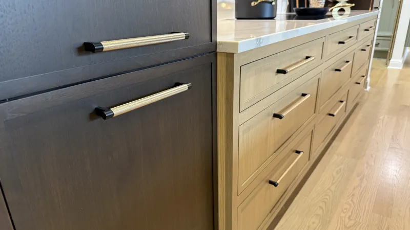 Modern kitchen cabinets with two-tone wood finish and brass handles under a white countertop on hardwood floor.