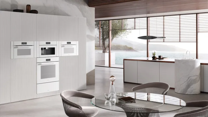 Modern kitchen with built-in white appliances, glass dining table, and large windows overlooking water and mountains.