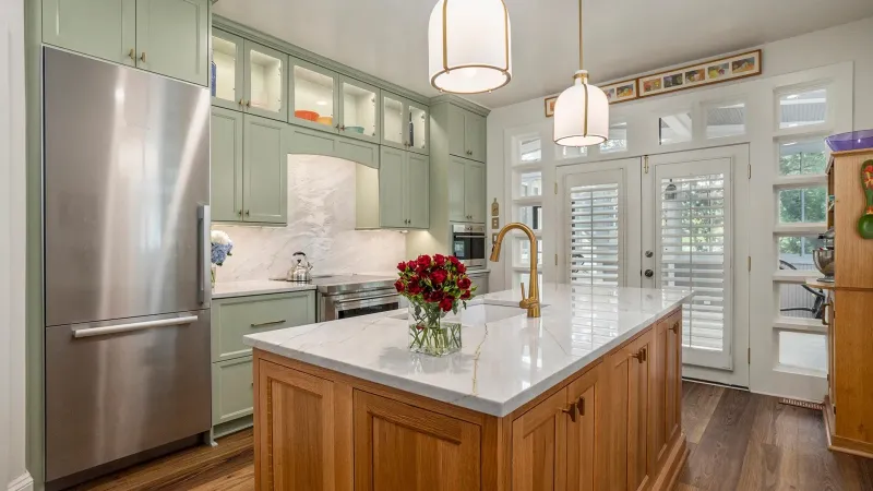Modern kitchen with green cabinets, wooden island, marble countertops, pendant lights, and stainless steel refrigerator.