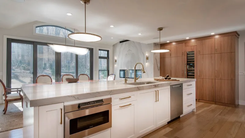 Modern kitchen with white marble island, brass fixtures, wood cabinets, stainless appliances, and large windows.