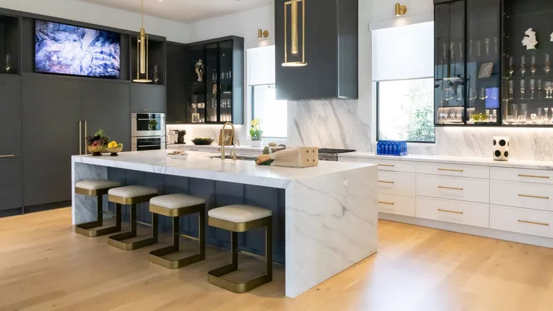Modern kitchen with marble island, black and white cabinetry, gold fixtures, and wooden flooring under natural light.