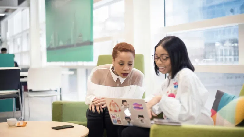 Two women collaborating and discussing work on a laptop in a bright modern office lounge area.