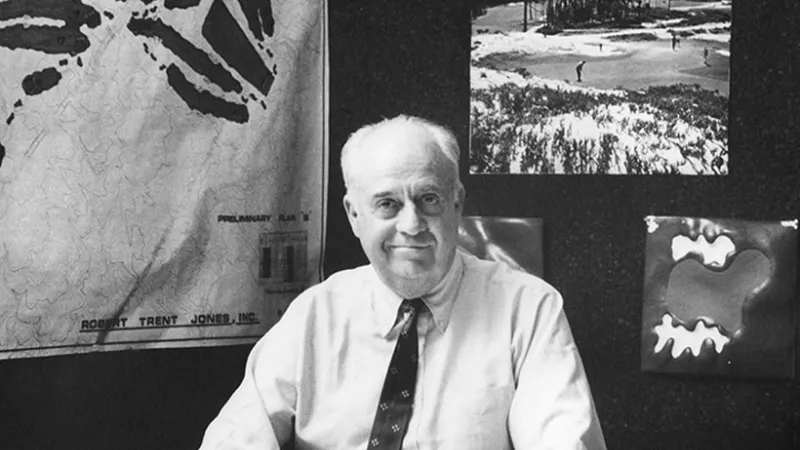 Black and white photo of a man sitting at a desk with maps and golf course plans on the wall behind him