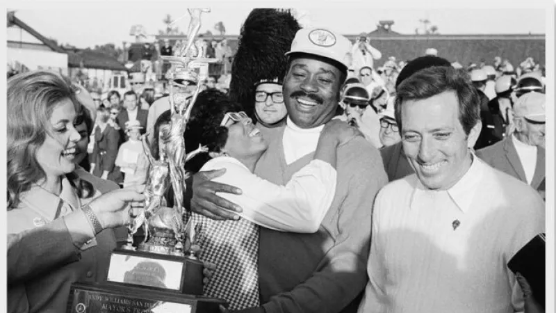 Group celebrating with a tall trophy, smiling and hugging, in an outdoor event with a crowd in the background