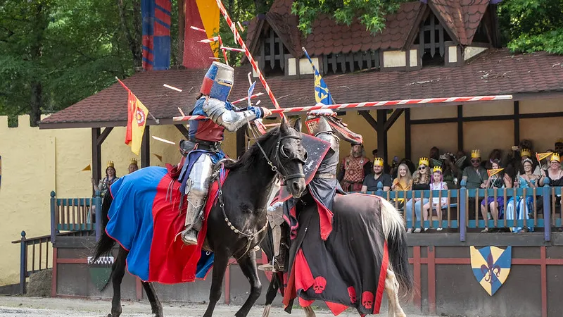 Two knights jousting on horseback during a medieval festival with spectators in period costumes and banners.