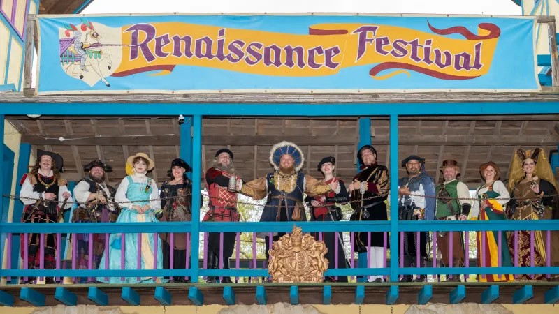 Group of people dressed in Renaissance costumes posing on balcony under Renaissance Festival banner.
