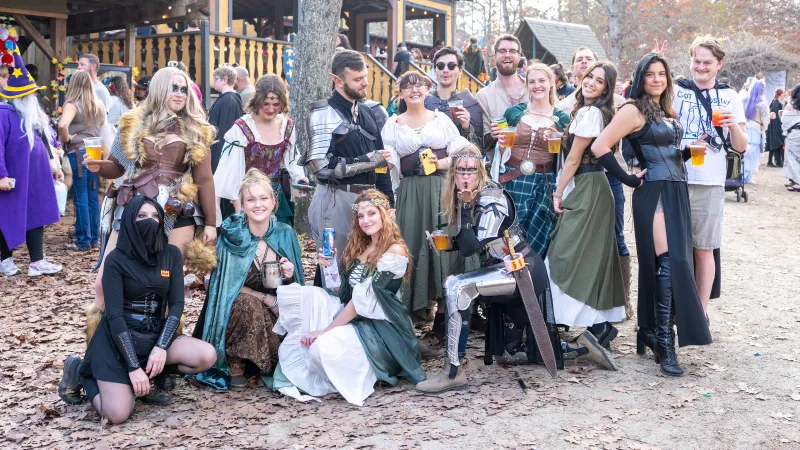 Group of people in medieval and fantasy costumes posing outdoors at a renaissance fair with drinks in hand.