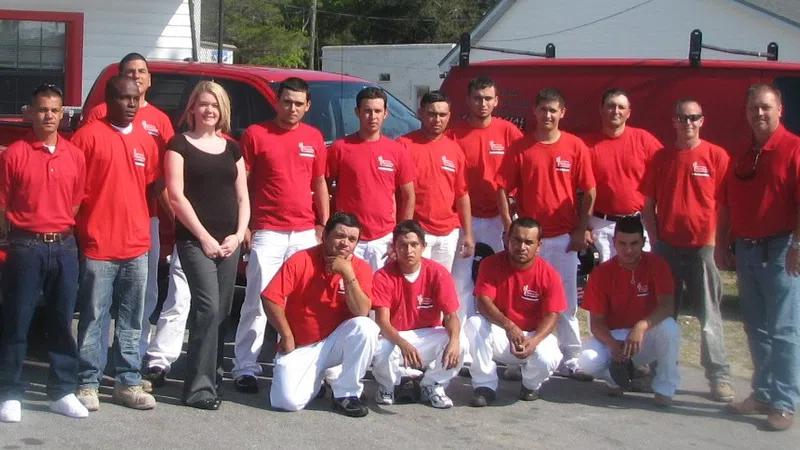 Group of 15 workers in red shirts and white pants posing outdoors near red trucks on a sunny day.