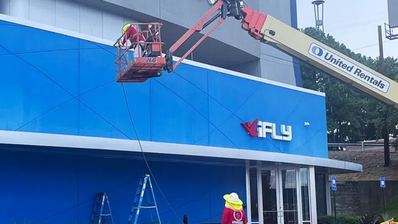 Aerial lift workers install or maintain the blue iFLY signage on a building exterior using cranes on a sunny day.