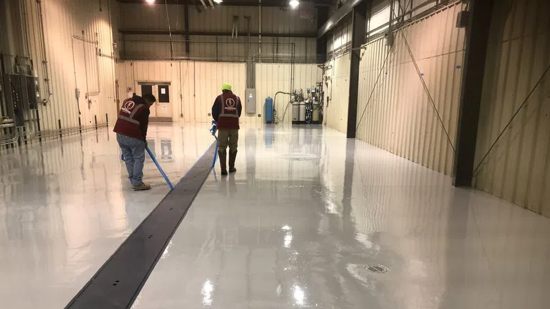 Two workers applying epoxy coating on a large industrial warehouse floor with high ceilings and metal walls.