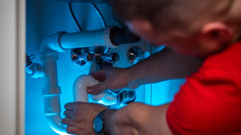 Plumber fixing white PVC pipes under sink illuminated by blue light, wearing red shirt and watch.