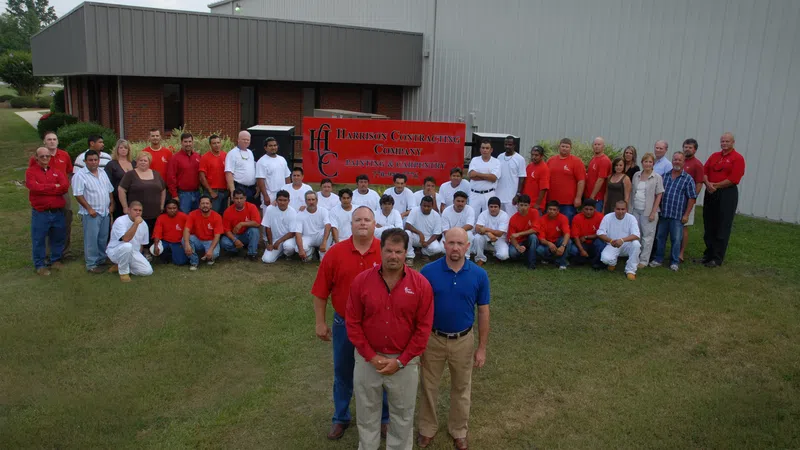 Group photo of Harrison Contracting Company staff posed outdoors on grass near building and company sign.