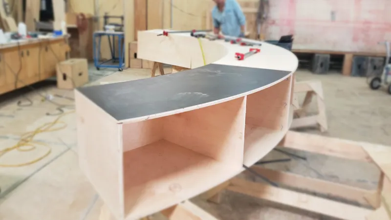 Curved wooden shelving unit under construction in a workshop with tools and blurred worker in background