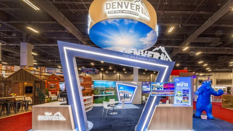 Modern Denver tourism booth with illuminated geometric frame, digital displays, and blue bear sculpture at expo