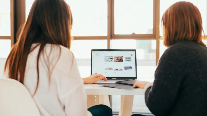 Two women browsing products on a laptop at a white table near a sunlit window in a modern office.