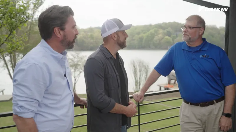 Three men engaged in conversation on a balcony overlooking a lake with trees in the background on a cloudy day