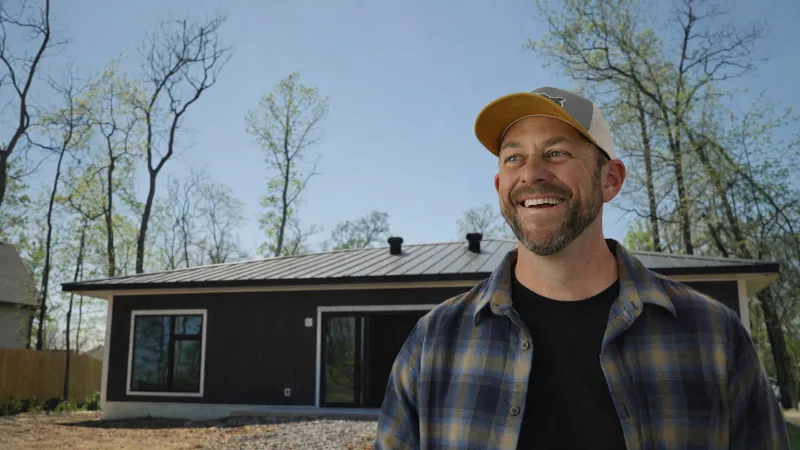 Smiling man in plaid shirt and cap stands in front of a modern black house with trees and clear sky.