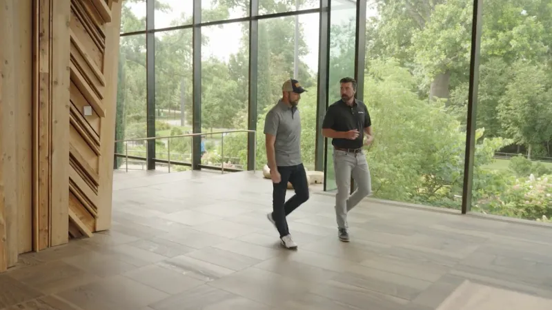 Two men walking and talking inside a modern building with large glass windows overlooking green trees.