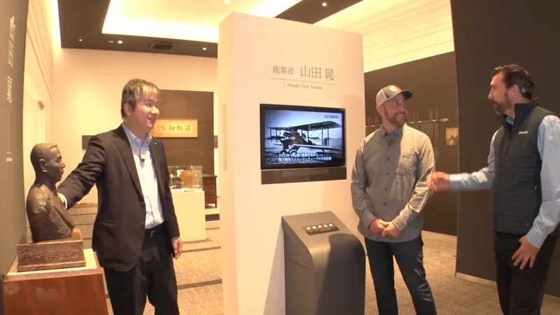 Three men in a museum exhibit discussing a bronze bust and a display about founder Akin Yamada.