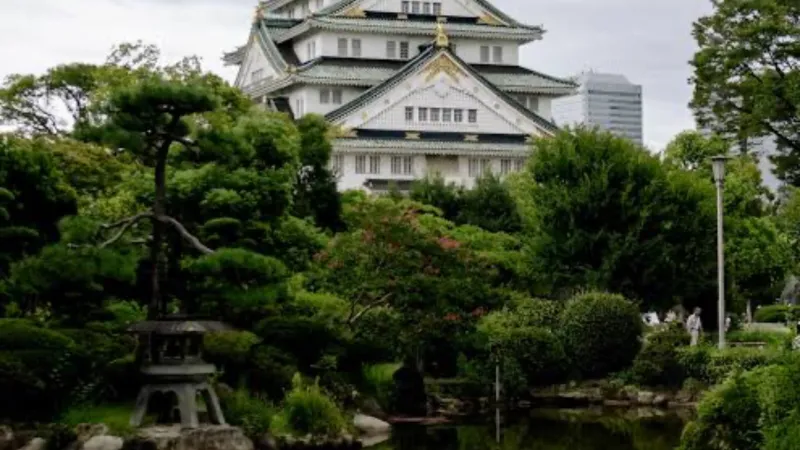 Osaka Castle reflected in a serene pond surrounded by lush green trees and garden under cloudy sky.