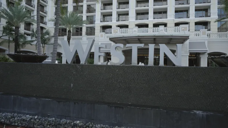 Westin hotel sign in front of a luxury building with palm trees and water features under daylight.