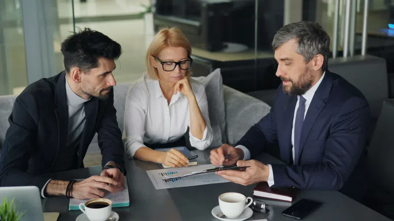 Three business professionals discussing charts and data during a meeting with coffee cups on the table