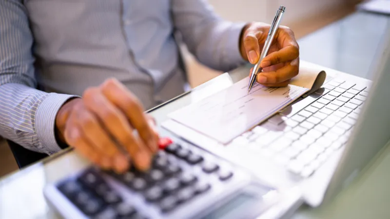 Person using calculator and writing on a checkbook at a desk with keyboard in an office setting.