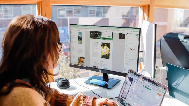 Person working on a laptop and desktop computer at a bright desk near windows with wooden frames.
