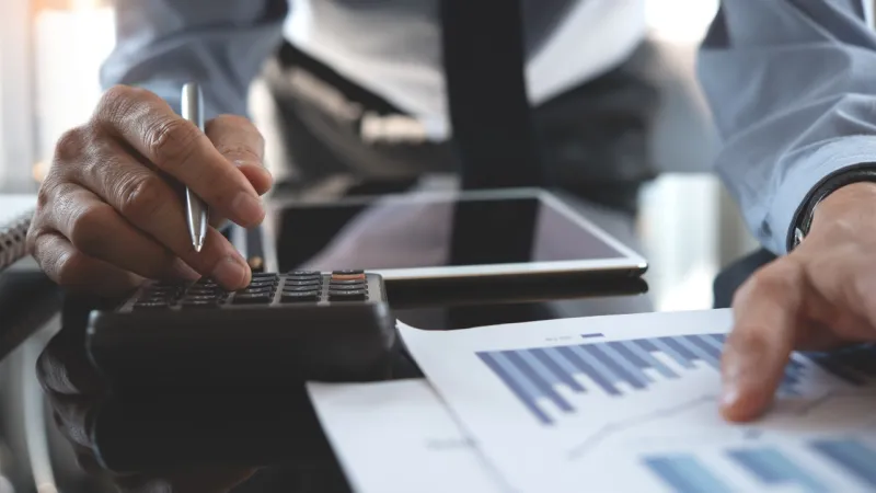 Person using a calculator and reviewing financial charts on paper with a tablet nearby on a desk.