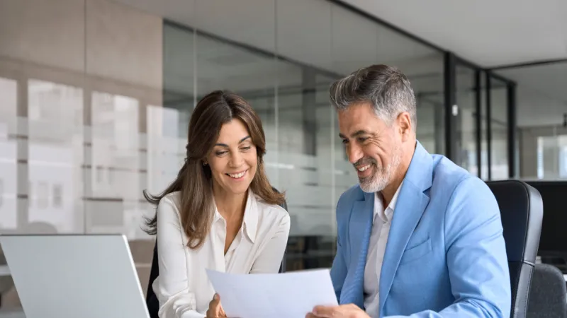 Smiling business colleagues review documents together at a modern office desk with a laptop and papers.