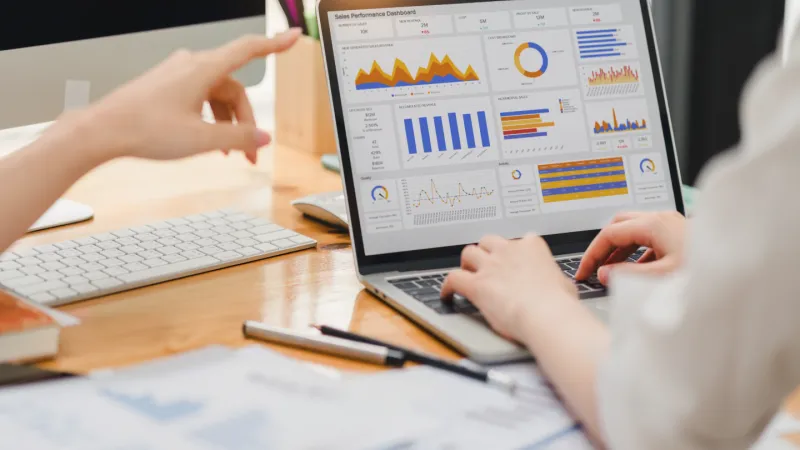 Two people analyzing sales performance charts and graphs on a laptop screen at a wooden desk.