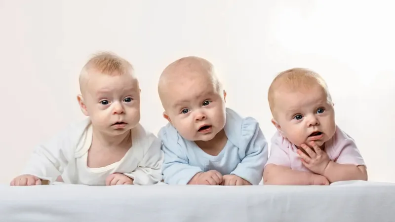Three curious babies lying on their stomachs on a white surface wearing pastel-colored clothes against a light background