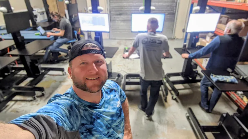 Man taking a selfie in a warehouse office with three coworkers working at standing desks and computer screens.