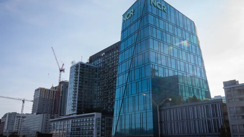 Modern glass office building with NCR logo in urban cityscape with cranes and traffic under clear sky.
