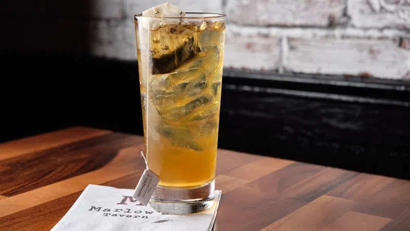 Tall glass of iced tea with tea bag on wooden table and Mason Tavern napkin against a brick wall.