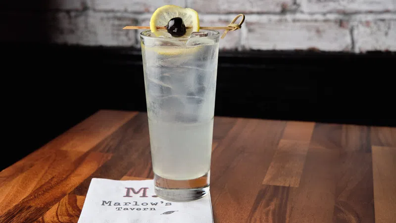 Tall glass of clear cocktail with ice, lemon slice, and cherry garnish on wooden table with Marlow's Tavern napkin