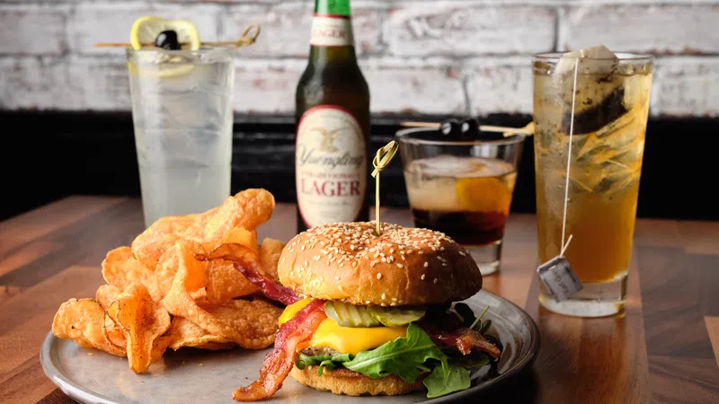 Cheeseburger with bacon, lettuce, pickles, potato chips, and drinks on a wooden table with brick wall background