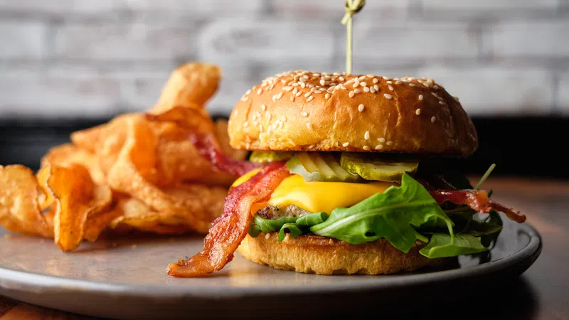 Juicy cheeseburger with bacon, pickles, and greens on sesame bun served with crispy potato chips on a gray plate.